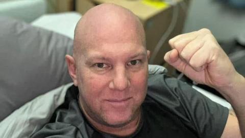 A man wearing a grey T-shirt sits in a bed looking up at the camera. He is punching the air with his left fist.