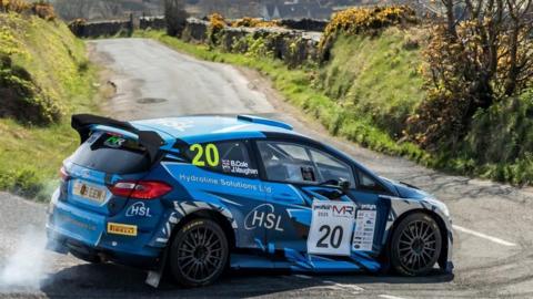 A blue rally car with the number 20 on the window rounds a bend on a country road.