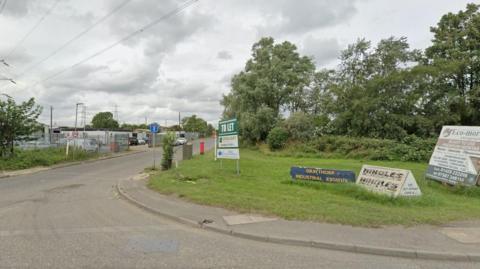 The entrance to Graythorp Industrial Estate. A blue sign with the name of the estate in yellow lettering stands in the grass by the side of the road. A number of other signs advertise businesses. In the background metal fencing stands around some of the warehouses.