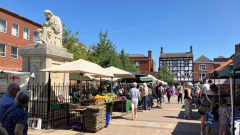 Lichfield town centre, with market stalls set up and people milling around. There is a clear blue sky, and a number of buildings surround the area where the market is being held.