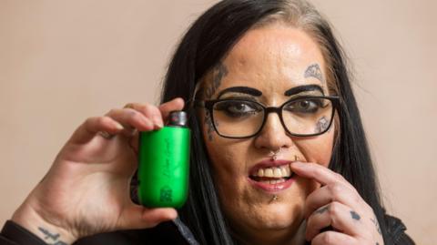 A woman holds a green vape up between her thumb and index finger beside her face while holding back her upper like with her other index finger. she has long straight black hair dark rimmed glasses, facial jewellry on her nose and lip and small tattoos on her cheeks and forehead 
