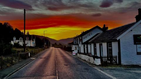 An orange and yellow sunset illuminates the street of the Inn. The road runs down the middle of the picture as the hotel sits to the right. Its a white, quaint building. It looks like a cottage and has the letters hotel printed on the side. 