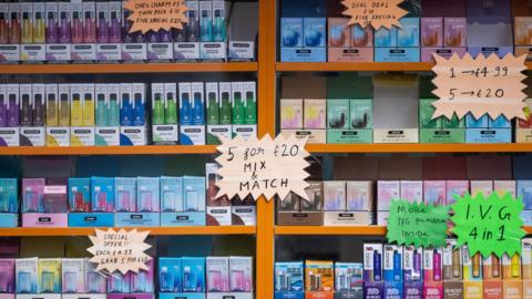 Bright coloured vape packaging on a shelf in a shop. Signs display offers like '5 for £20'
