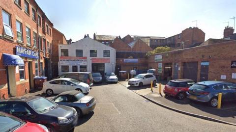 A short road in Northampton, showing lots of parked cars, red brick buildings, businesses and roof tops. 