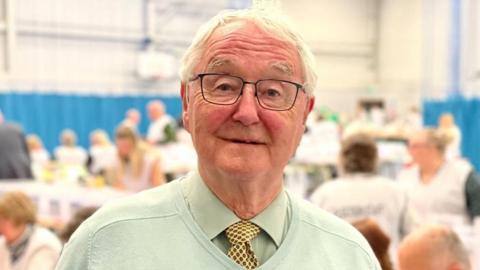 A man with white hair, glasses and a pale green jumper in a crowded room with the blurred shapes of people behind him