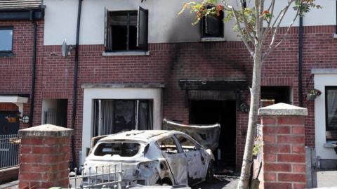 A burnt-out car in the driveway of a house which has black smoke damage on its front wall. A gate is also damaged.