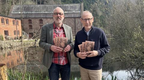 Two men are holding brown paper books while standing up looking at the camera. Behind them are buildings and a lake.
