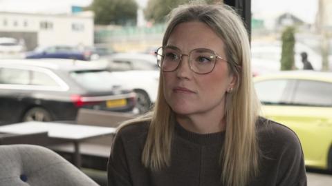 A woman looks slightly to the side, she does not smile. She has blonde hair and wears glasses, and a brown knit jumper. She is sitting inside a café and there's a big window behind her.  There is a car park in the background.