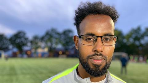 Yousuf has black hair and a goatee beard. He is wearing black framed square glasses and a grey top with yellow fluorescent stripes along the shoulders. Behind him is a field and trees in soft focus. 