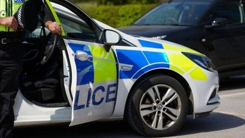 A generic police car parked on a road. A police officer holds the driver's door open with one hand and hold their police hat in the other.