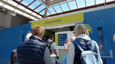 Passengers queuing to go through departures at Jersey's ferry terminal. The photo is taken over the shoulder of the passengers.