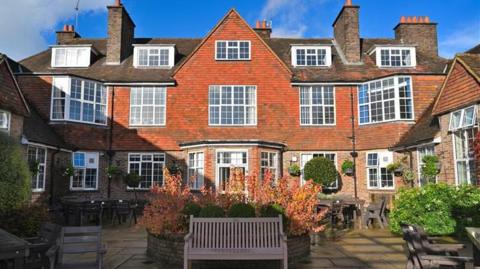 A large building which has a red tiled facade, several chimneys and a courtyard with assorted tables and chairs outside.
