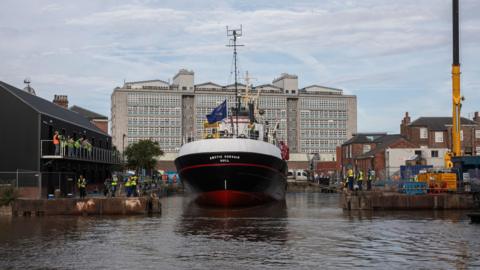 The trawler Arctic Corsair - a black-hulled boat with white upper works - enters a dock from the waters of the River Hull. People watch from the banks and the balcony of a black two-storey building. The skyline is dominated by a large concrete and glass building.
