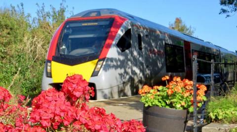 A Greater Anglia train, which is grey with red and yellow stripes at its front, is drawn up alongside a station platform. There are red and orange flowers in the foreground. It is a bright, sunny day and the sky is blue.