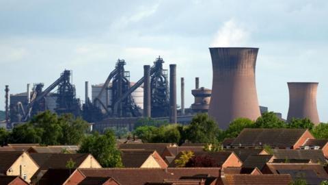Brown roofs and leafy green trees sit in front of the squat brown blast furnace smokestacks and industrial pipes and silos of the British Steel plant in Scunthorpe.