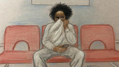 Court sketch showing Axel Rudakubana sat in the middle of a row of three red chairs. He's wearing a grey tracksuit, holding an item in his left hand and his right hand is holding his sweater onto his face - hiding his nose and mouth