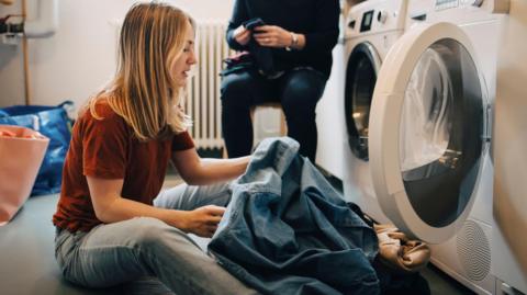 A young blonde woman wearing light blue jeans and a burnt orange t shirt sits on the floor in front of a tumble drier taking her clothes out of it