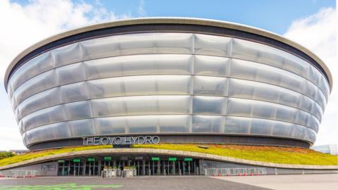 The Ovo Hydro is a tall, round silver building which looks like several large silver bands piled on top of one another. The entrance has several doorways and a sign above the entrance reads THE OVO HYDRO. The entrance has a small band of grass on top of it which snakes around the building. The land approaching the entrance is smooth grey pavement.