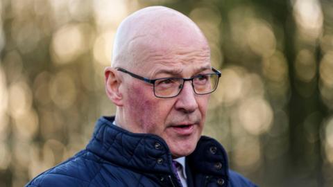 John Swinney, who is bald, is wearing black glasses and a navy padded jacket. He appears to be outside and mid-speech.