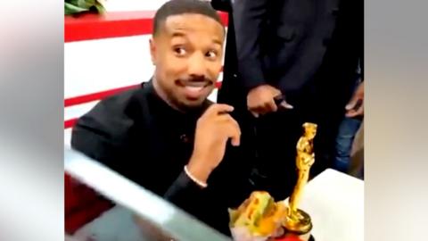 Michael B Jordan smiles at an In-N-Out Burger restaurant as he holds a burger. His Oscar sits on the table next to him.