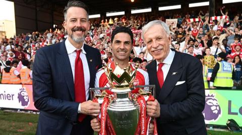 Josh Kroenke, Mikel Arteta and Stan Kroenke posed with the Premier League trophy, standing in front of a stand of Arsenal supporters who are largely dressed in red and white. Josh and Stan Kroenke wear dark suits, white shirts and a red tie each, while Arteta is in a red Arsenal shirt