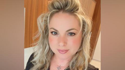 A woman with long, wavy blonde hair. She is wearing a black V-neck T-shirt and has a tattoo of a sparkle at the top of her chest.