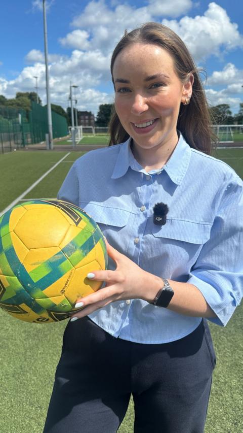 A smiling woman in a light blue shirt holds a yellow football. She is standing on a football pitch.