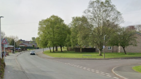 A car is driving along Unicorn Road before a junction. Trees and grass surround some housing on the right side of the street