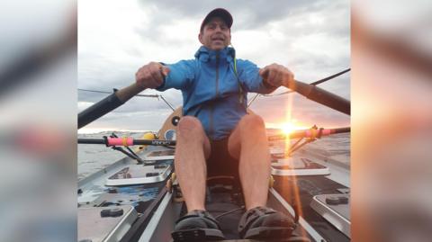 Billy Taylor seated in a rowing boat on the ocean and facing the camera. He has a blue jacket and cap on and is holding an oar in each hand. The sun is setting behind him.