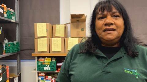 Ruth Masih in a green polo shirt with a Farnborough Foodbank logo standing in front of shelves with food items in boxes