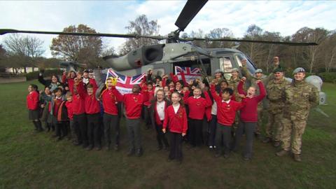 Schoolchildren and military personnel stood on a field in front of a helicopter cheering.