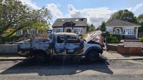 A burnt out silver pick up truck on the road in front of a bungalow with a damaged roof with holes in.