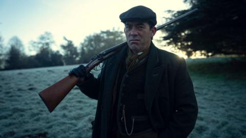 A man in a 1920s - 1930s style suit is standing outside in a frosted field with a shotgun held over his shoulder. He is wearing a peaked flat cap, woollen overcoat, woollen waistcoat and heavy scarf in a golf colour. There are trees visible in the middle distance.