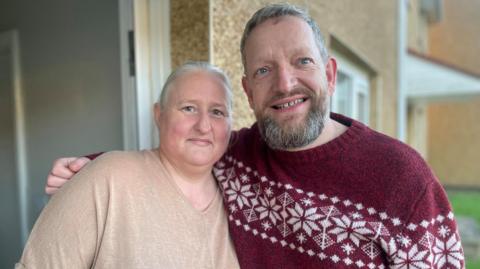 A woman with grey hair tied back smiles to the camera shyly whilst a bearded man with a Christmas jumper puts his arm around her