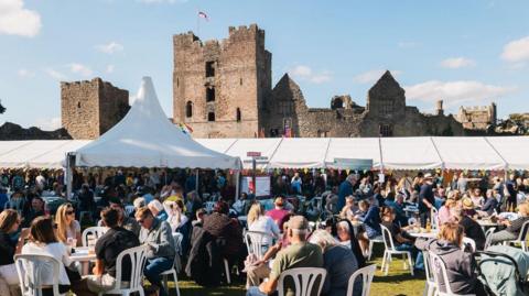 Crowds of people, some of them seated with marquees in the distance and a castle behind them