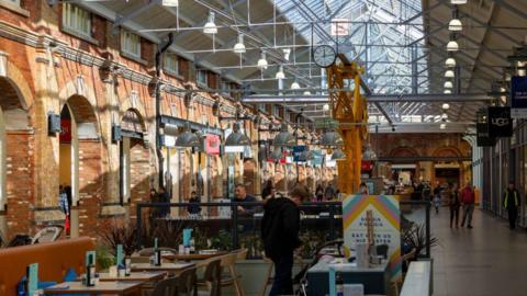 Restored former railway shed, now turned into a modern shopping centre, with glass and metal roof, with brick-built arches running the length. Different shops are nestled into each of the arches. Most cannot be read, but one reads UGG, some restaurant tables can be seen in the foreground, and a yellow crane from the centre's days as a railway yard is in the middle of the lines of shops