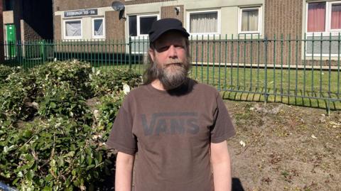 Andy stands in front of the flats. He wears a brown t-shirt with a black Vans logo and a black cap. He has a medium length brown and grey beard and long brown and grey hair,