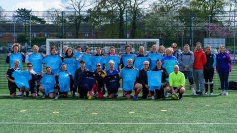 Women's walking football team stood holding blue shirts in front of a goal on a football pitch. There are houses and trees in the background.