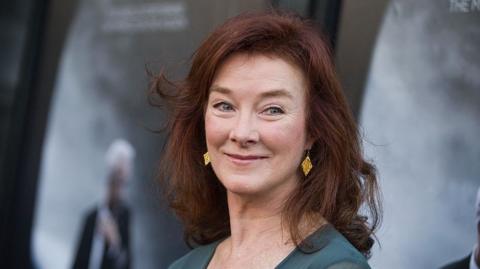 Valerie Mahaffey is pictured in front of a grey background. She has red hair and is wearing a green dress and gold earrings. She has blue eyes.