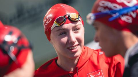 Siena Stephens (in focus), wearing red swimming hat, gold and white swimming goggles and red training top with toggles, speaks to other triathlon competitors