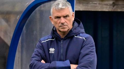 John Askey with his arms crossed while stood in the dugout