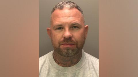 A police custody photograph of Michael Watling. He is wearing a grey jumper. Watling has short, greying hair and a short beard. His neck is also covered in tattoos.