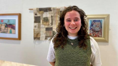 A woman wearing a white and green top stands in front of pieces of art. She is smiling at the camera. Behind her lies two paintings, as well as a piece of patchwork.
