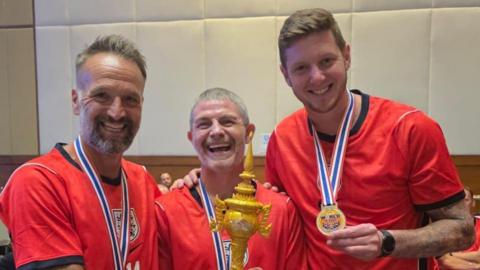 Karl Duguid, Jamie Cureton and Greg Halford all laughing or smiling while holding their winners' medals. Cureton is holding a large golden trophy. They are all wearing a red England top.