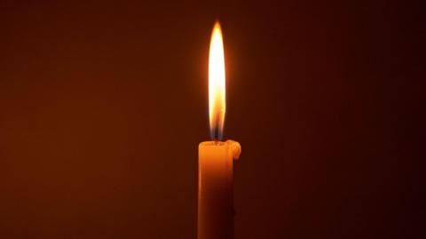 A single-lit candle casting a warm and serene glow in a dark background. There is a tall flame and some wax dripping down the candle.