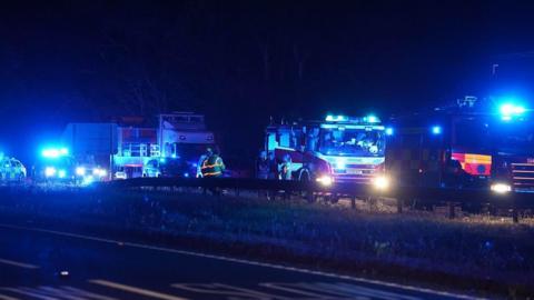 Emergency service vehicles, including fire engines, police vehicles and lorries, are stopped on the A27 at night. Blue emergency lights illuminate the scene while several people stand near the central reservation.