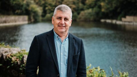 Helier Smith standing in front of a body of water and waist-height stone wall, looking directly into the camera, wearing a smart dark blue jacket with lighter blue button up shirt below