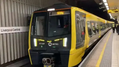 A yellow train with the destination Kirkby at Liverpool Central station platform. Staff in hi-vis vests and a passenger are seen in the background.