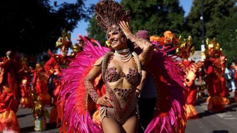 A female dancer wearing bright pink costume, with lots of dancers in bright costumes behind her.