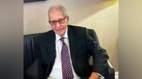 An elderly man with thinning grey hair and glasses, wearing a white pinstripe shirt, colourful tie and a dark blazer sits on a seat looking into the camera.
